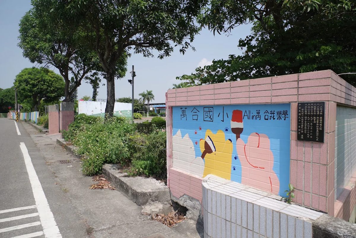 萬合國小校門旁的彩繪圍牆：粉紅色磚牆上一片藍色彩繪，寫著「萬合國小 All in 萬合我樂學」，畫著彩色的動物與一支冰棒，右側嵌著一塊黑色石碑，牆外是一排老樹與鄉間道路 The painted wall beside the Wanhe gate — a blue panel set into pink brick reading 萬合國小 All in 萬合我樂學, with coloured animal shapes and an ice lolly painted across it, a black stone tablet set in at the right, and old trees and a country road outside