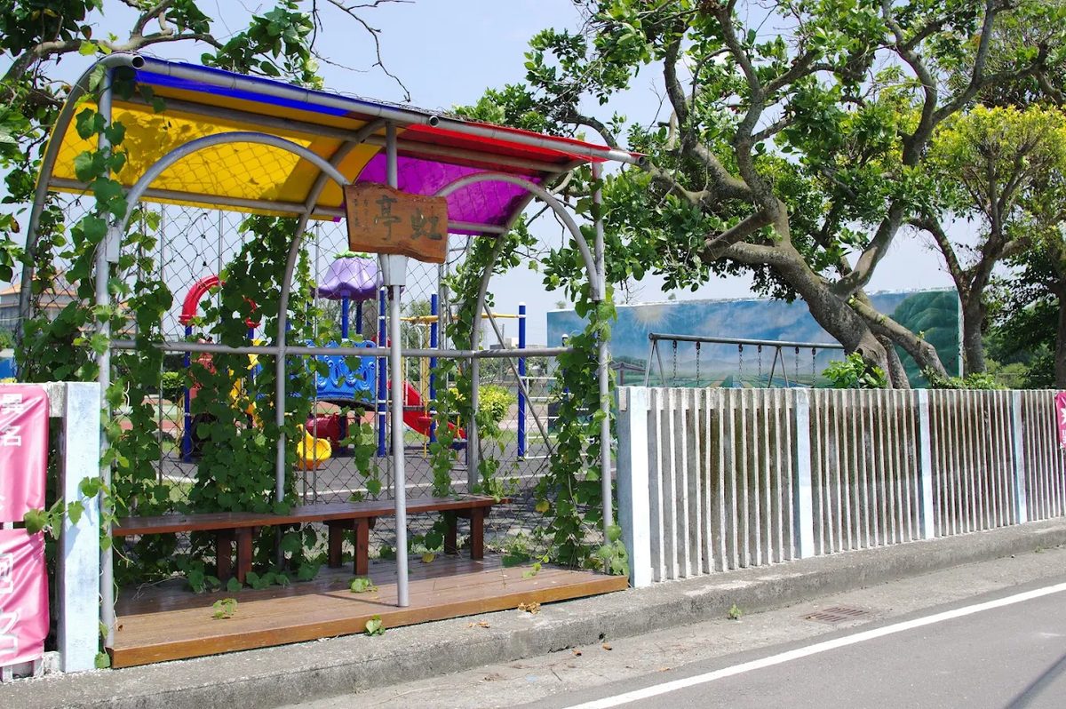 萬合國小圍牆邊的等候亭「虹亭」：弧形鋼架撐起黃、紫、藍、紅的彩色頂棚，藤蔓爬滿兩側，亭內是木地板與長木椅，木牌上寫著「虹亭」二字，亭外可以看見遊具與白色欄杆 The 虹亭 waiting shelter along the Wanhe boundary wall — a curved steel frame carrying a canopy in yellow, purple, blue and red, vines climbing both sides, a timber deck and bench inside, a wooden sign reading 虹亭, and the play equipment and white railings visible beyond