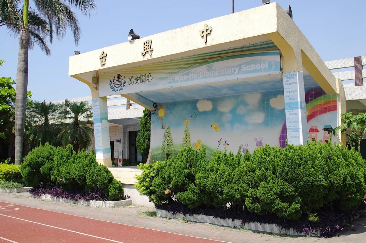 萬合國小司令台側面：米黃色台體上方由右至左寫著「中興台」，前緣印著校徽、「萬合國民小學」與「Wan-He Elementary School」，背板是藍天白雲、小樹與彩虹的彩繪，台前是一排修剪整齊的圓球狀灌木與紫色地被 The stage at Wanhe seen from the side — the cream structure with 中興台 across the top, the crest and the names 萬合國民小學 and Wan-He Elementary School along its front edge, a painted backdrop of blue sky, small trees and a rainbow, and a row of ball-clipped shrubs and purple ground cover in front