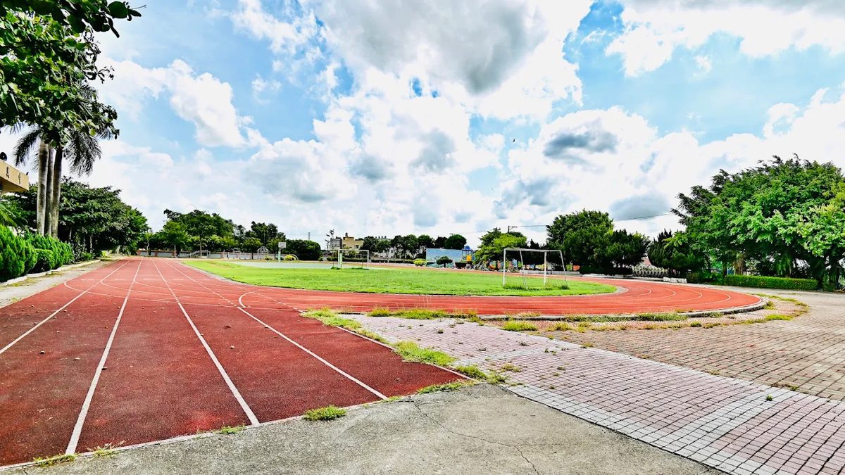 萬合國小的操場：紅色 PU 跑道環繞一片綠色草地，遠處有籃球架、鞦韆與樹籬，天空藍白相間、白雲成堆 The sports field at Wanhe — a red synthetic track curving around a green infield, with a basketball hoop, swings and clipped hedges at the far end under a blue sky banked with white cloud