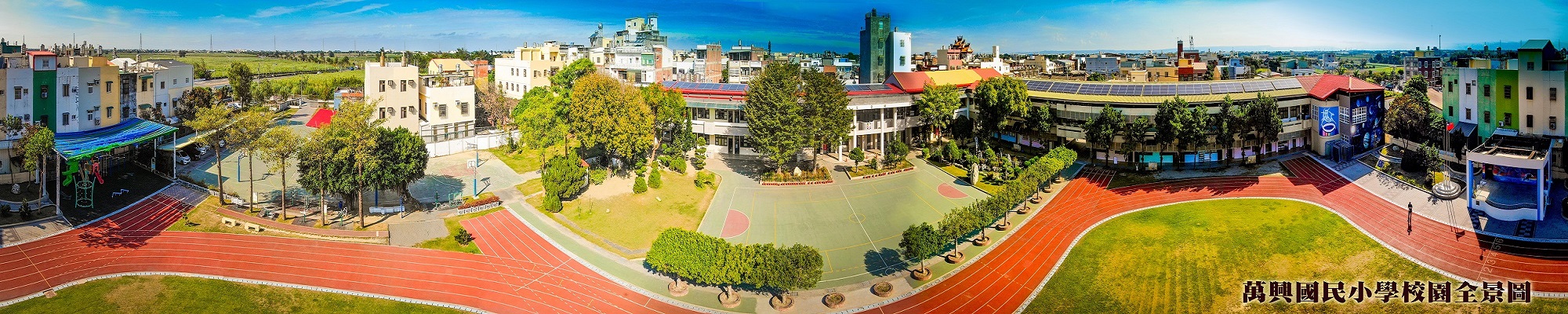Aerial panorama of Wansing Elementary School — a U-shaped two-story white campus surrounding a red running track, framed by paddy fields and village houses
