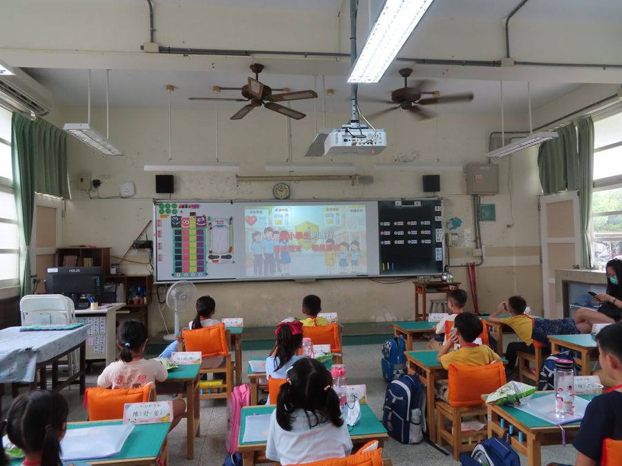 小一新生第一天在教室裡，桌上放著小書包，老師在台前 New first-graders on their first morning in the classroom, little backpacks on the desks