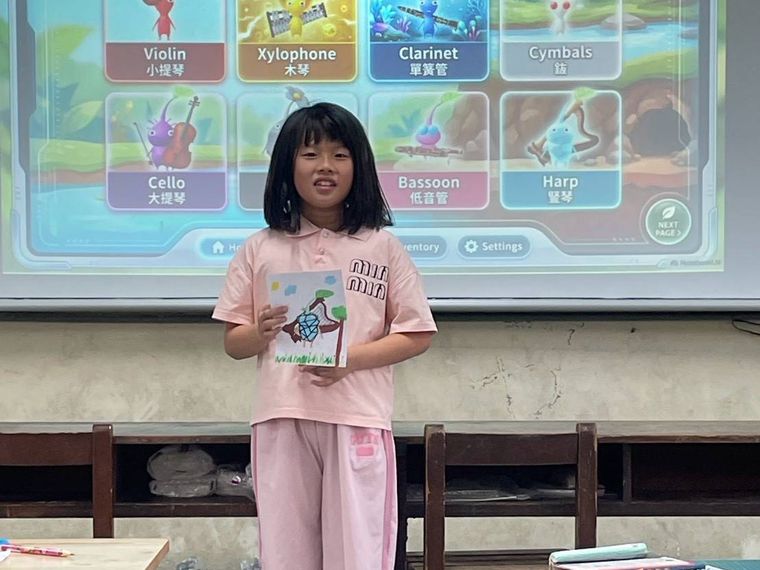 源泉國小英語社學生站在教室前，以英語介紹樂器主題，投影幕上有小提琴、豎笛等單字 A Yuanchyuan English club student giving an English presentation on musical instruments at the front of the class
