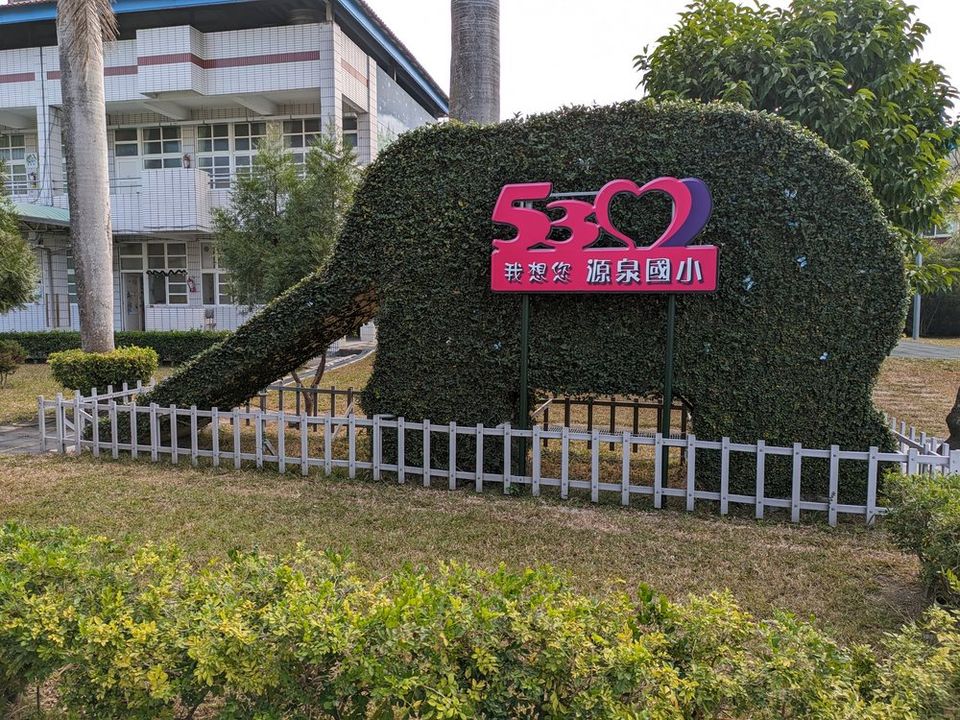 源泉國小庭園中一座修剪成大象形狀的綠籬，前方立著粉紅色「530 我想您 源泉國小」看板 An elephant-shaped topiary hedge in the Yuanchyuan garden, with a pink '530 · we miss you · Yuanchyuan' sign in front