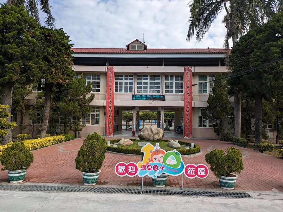 源泉國小主校舍正面，前方庭園中央立著「歡迎源泉國小100週年蒞臨」的彩色看板 The front of the Yuanchyuan main building, with a colourful '100th anniversary welcome' sign in the garden before it