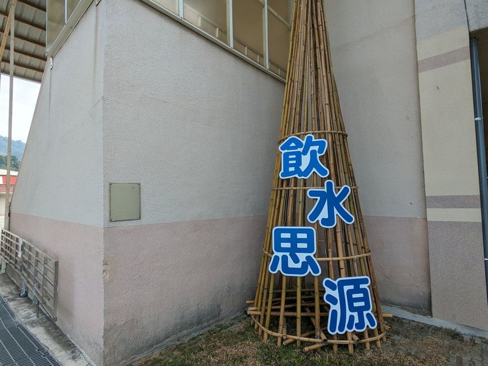 源泉國小牆邊一座竹編圓錐裝置，前面立著藍色大字「飲水思源」 A woven-bamboo cone by a Yuanchyuan wall, with the large blue characters for 'remember the source of the water' in front