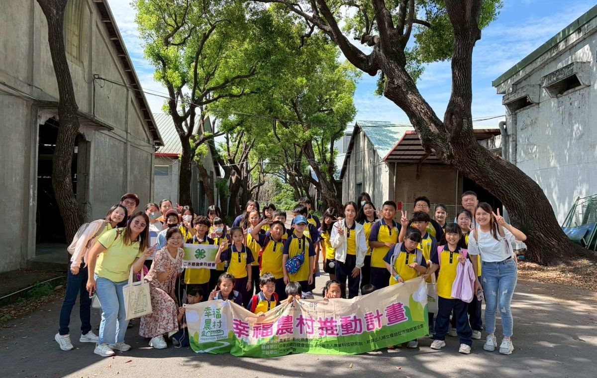 源泉國小師生在大樹下進行社區踏查，手持「幸福農村推動計畫」布條，藍天綠樹 Yuanchyuan students and teachers on a community field walk under big trees, holding a rural-community project banner