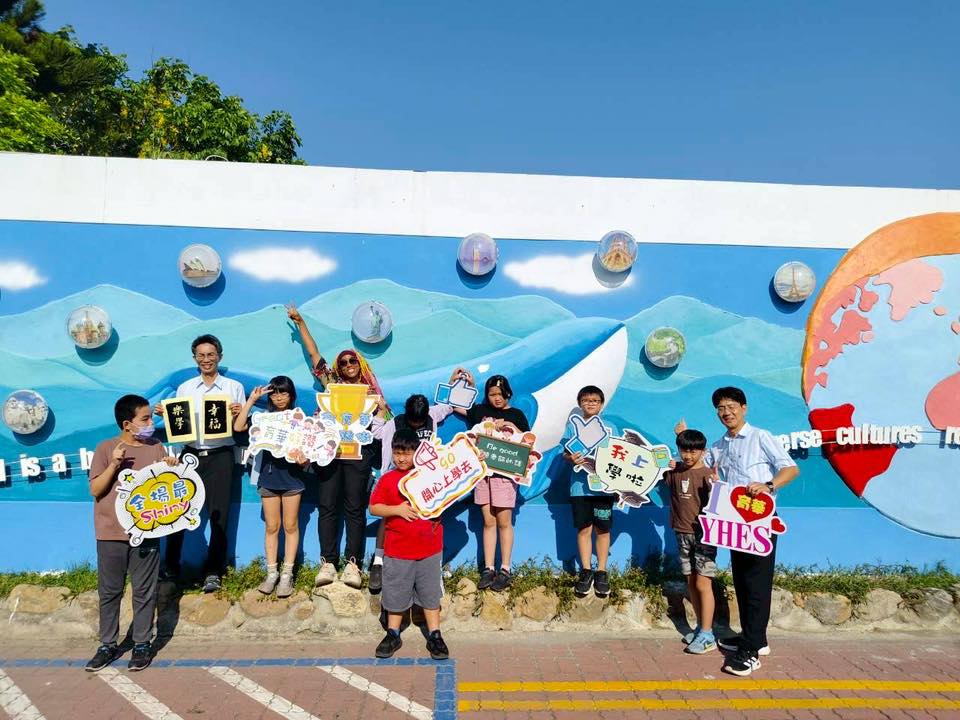 Students and Dom Jones pose in front of the hand-painted world-map mural holding craft signs