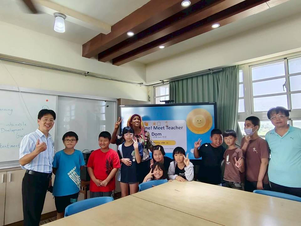 Students pose by a screen reading Welcome! Meet Teacher Dom in the classroom