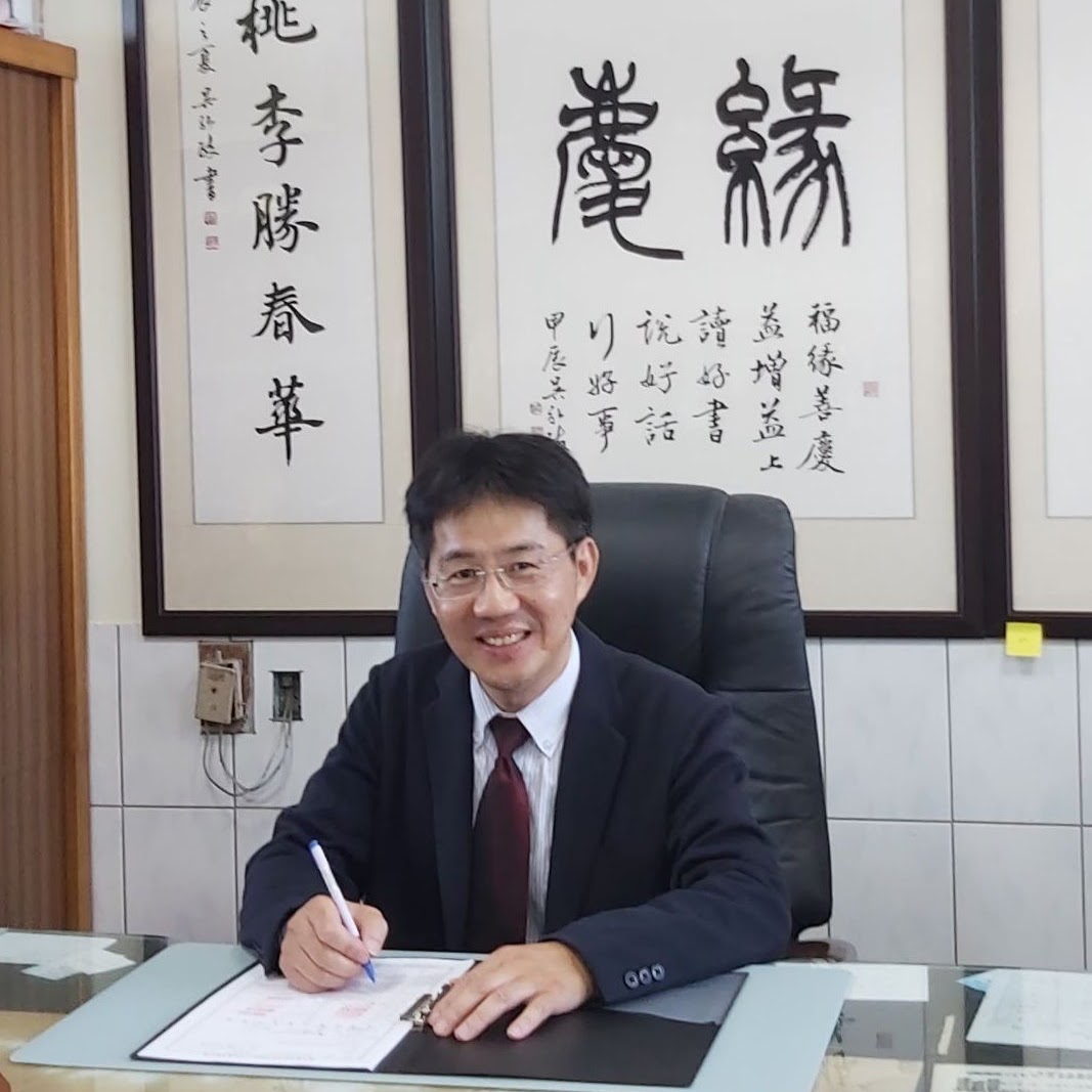 Principal Chen Chih-ming at his desk, with calligraphy reading 桃李勝春華 and 緣慶 hanging behind him
